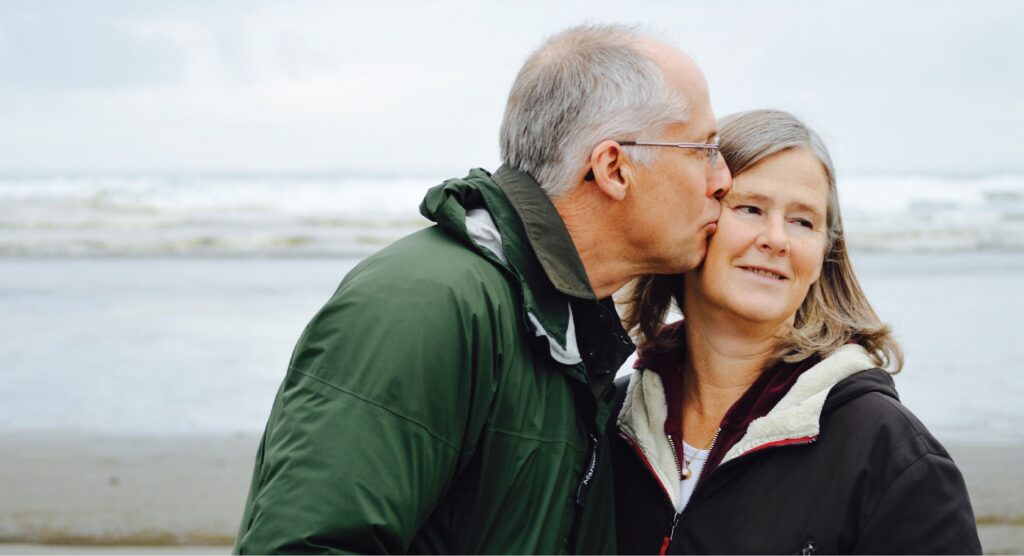 A senior man kissing his wife's cheek with a shoreline behind them.