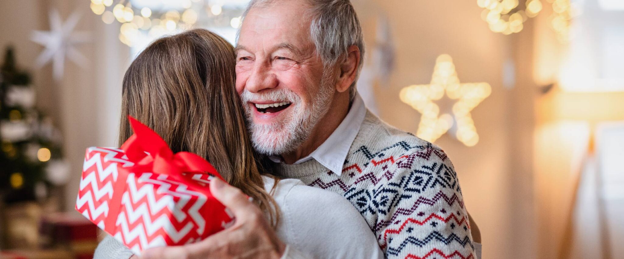 A senior man hugs his daughter while holding a red wrapped gift.