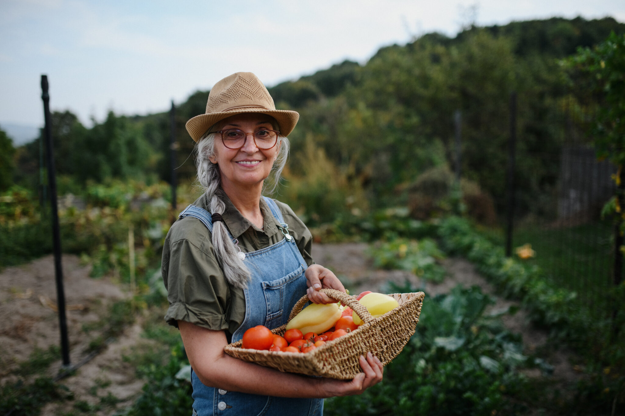 A senior woman in overalls holds a basket of produce in her outdoor garden.