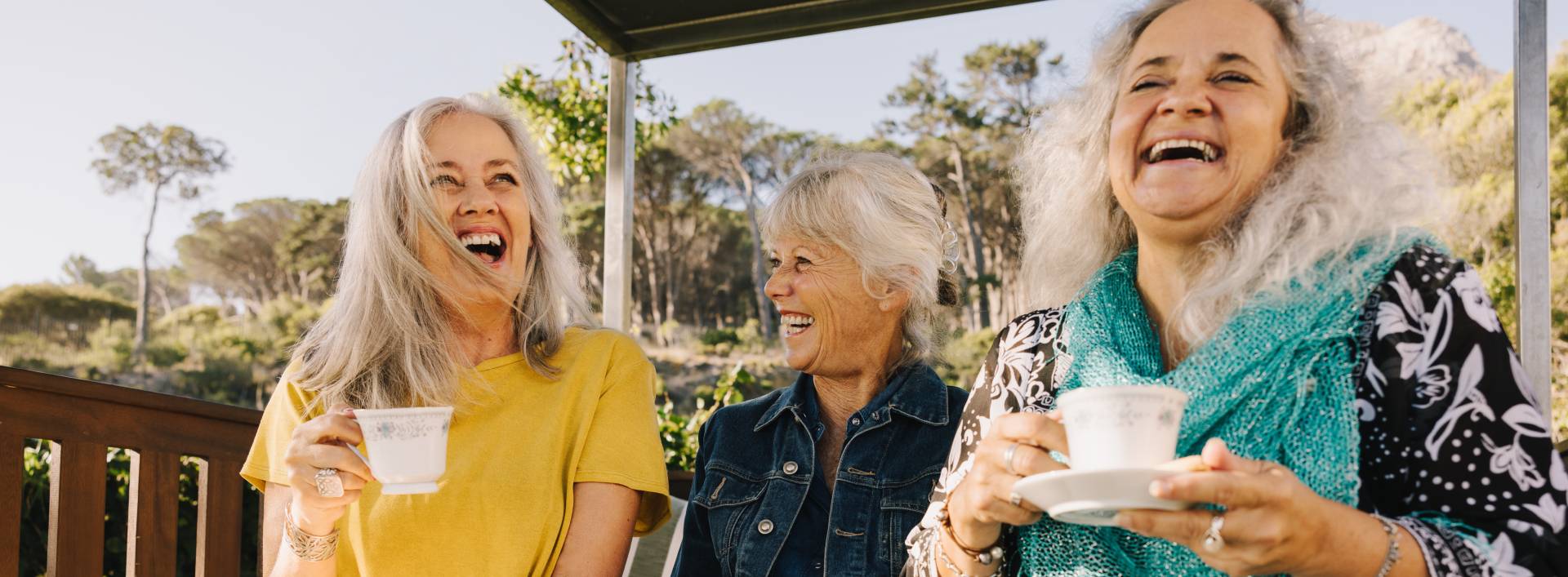 three senior ladies sitting on a bench outside drinking coffee and laughing together