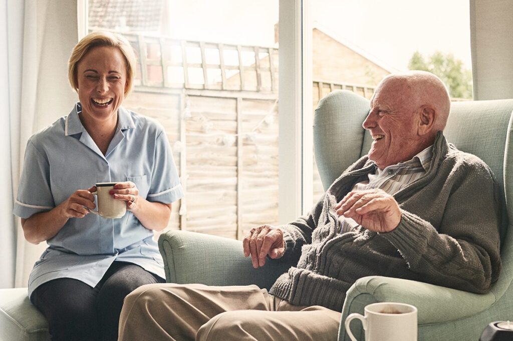 A senior man laughs with a nurse over coffee.