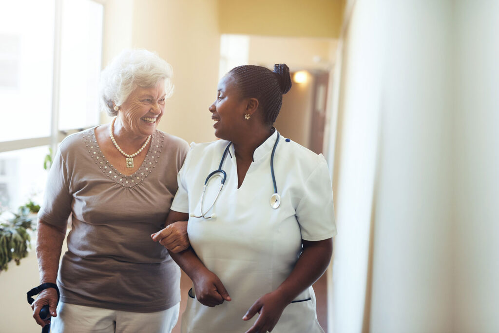 A senior woman laughing and walking with assistance from a nurse.