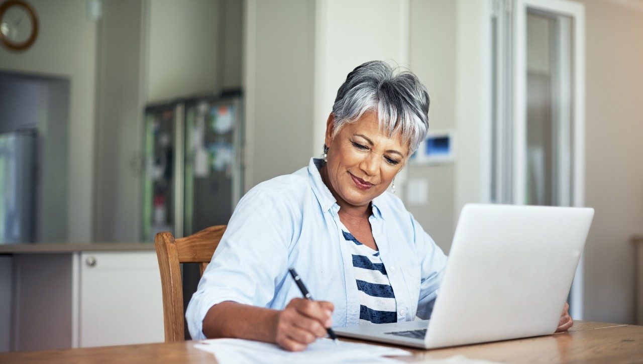 fvholl An older woman sits down at a table with her laptop and looks at the paperwork next to her