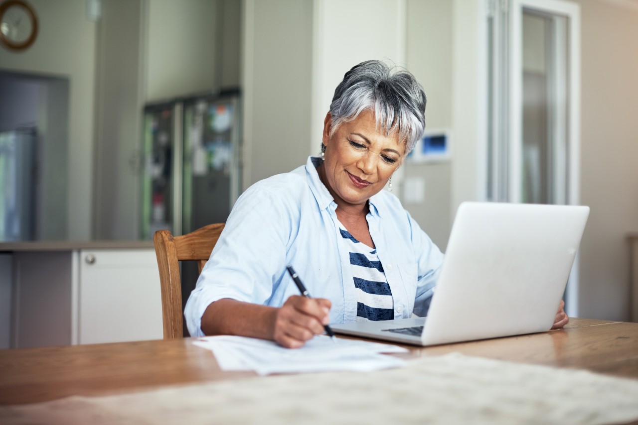 An older woman sits down at a table with her laptop and looks at the paperwork next to her