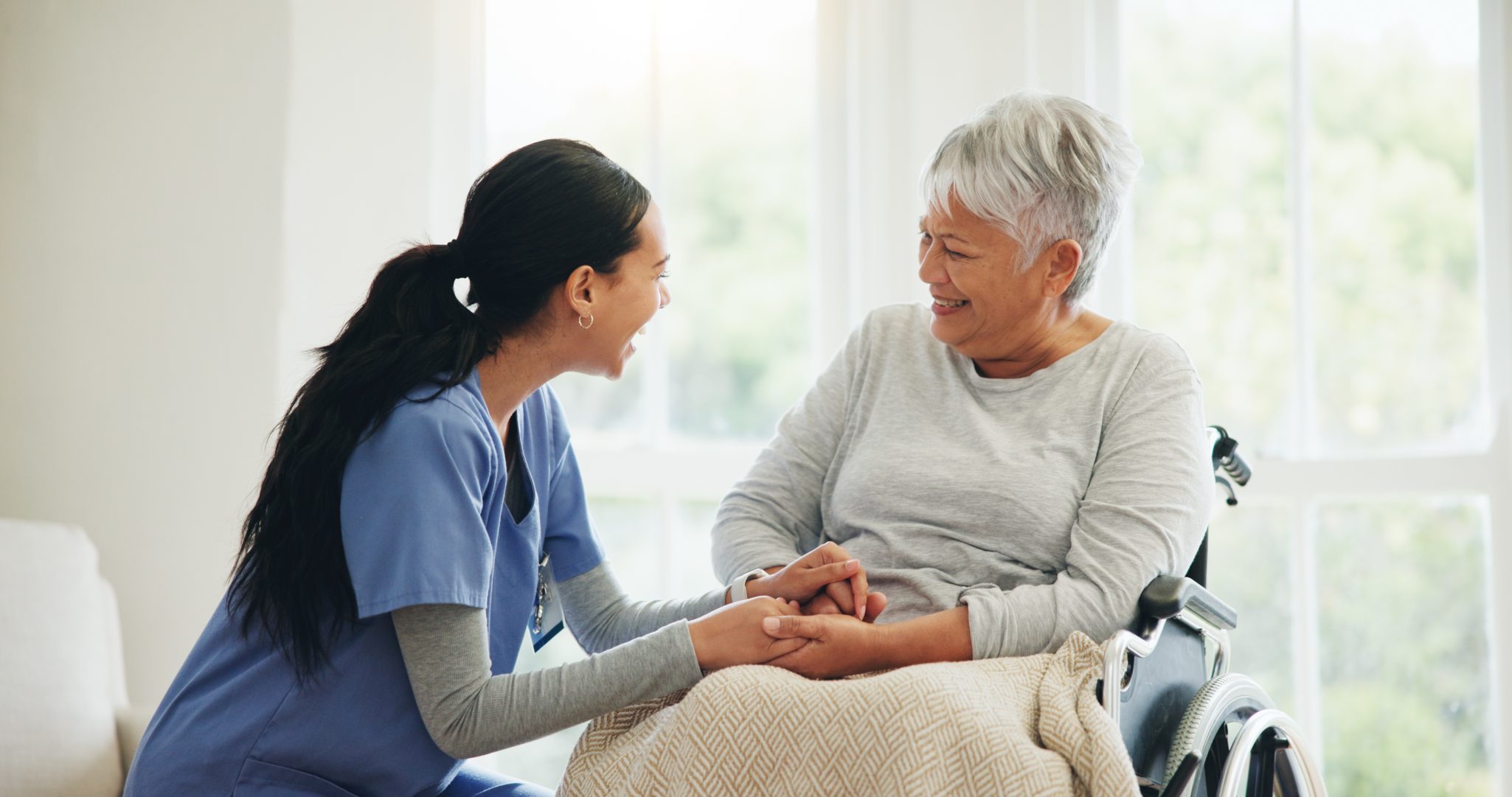 A senior woman in a wheelchair smiling at her caretaker who's holding her hands