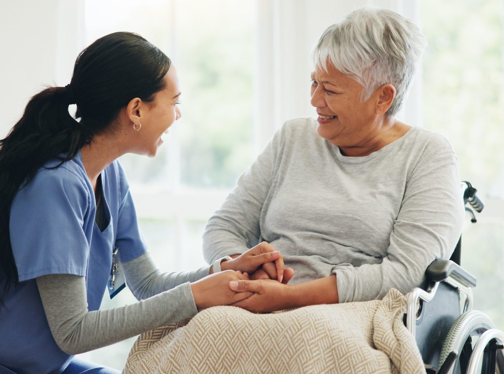 A senior woman in a wheelchair smiling at her caretaker who's holding her hands